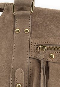 Détail d'un sac à main en suede marron avec quincaillerie dorée, accents de couture et une poche zippée ornée d'un gland décoratif.