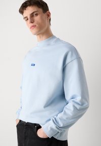 Felpa azzurra realizzata in materiale morbido, con un alto collo e una piccola toppa con logo blu sul petto. Vestibilità comoda con maniche lunghe.