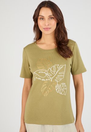 Frau trägt ein olivgrünes T-Shirt mit weißen und orangen tropischen Blattumrissen und dem Wort „summer“ in Kursivschrift auf der Vorderseite.