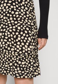 Svart klänning med ett krämfärgat oregelbundet prickmönster, en volangnederdel och ett mjukt tyg. En hand med en långärmad svart topp syns.