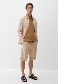 Jasno beżowa lniana marynarka z guzikami, brązowa teksturowana koszulka polo, beżowe szorty i czarne sandały. Prosty, codzienny design.