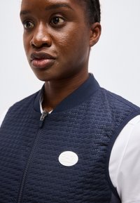 Μαύρο άτομο που φοράει μπλε ναυτικό αμάνικο μπουφάν με καπιτονέ επένδυση πάνω από λευκό πουκάμισο, κοιτάζοντας ελαφρώς προς τα αριστερά με ουδέτερη έκφραση.