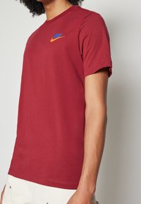 Czerwona bawełniana koszulka z okrągłym dekoltem, krótkimi rękawami oraz haftowanym niebiesko-pomarańczowym logo Nike na piersi. Prosty design, gładka tekstura.