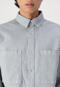 Lyseblå denimskjorte med krage, trykknap-lukning og to brystlommer. Har en blød tekstur og subtile sydetaljer.