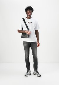 Camiseta blanca de manga corta con un logo, bolsa cruzada negra con texto en relieve, jeans ajustados gris, y zapatillas deportivas en blanco y negro.