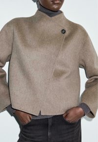 Cappotto in misto lana marrone con colletto alto, davanti asimmetrico e un singolo dettaglio con bottone nero, abbinato a un dolcevita grigio sotto.