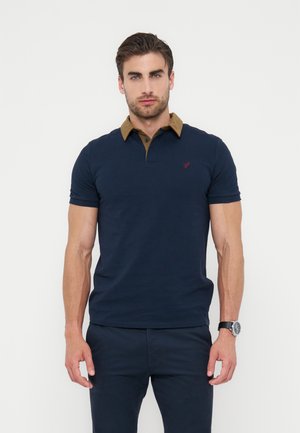 Mand med brunt hår iført navy polo-shirt med kontrasterende brun krave og navy bukser, stående mod en ensfarvet hvid baggrund.