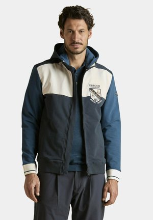 Uomo che indossa una giacca con cappuccio con zip nei colori blu navy, crema e blu, con il logo delle Frecce Tricolori sul petto sinistro, in piedi su sfondo bianco.