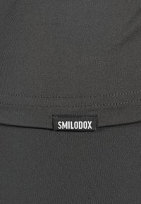 Zwart atletisch materiaal met een gladde textuur, met een zichtbaar "SMILODOX" etiket en stiksels langs de zoom.