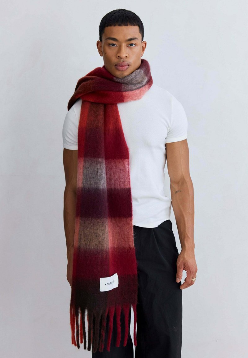 Écharpe à carreaux rouge et bordeaux fabriquée en tissu texturé, avec des extrémités à franges et une étiquette blanche portant la mention "ARCTIC." Portée avec une chemise blanche.