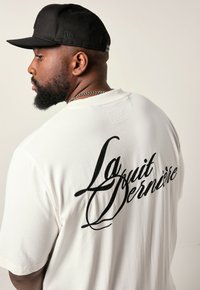 Man met een baard die een zwarte pet en een wit shirt draagt met de tekst "La Squit Bernesire" op de rug, kijkend naar links tegen een effen achtergrond.