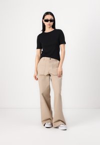 T-shirt nero a maniche corte abbinato a pantaloni beige a vita alta e palazzo, con sneakers bianco e nere; accentuato da occhiali da sole scuri.