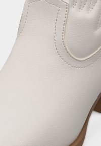 Κοντινό πλάνο λευκής δερμάτινης μπότες με ραμμένα διακοσμητικά σχέδια και ανοιχτόκαστο τακούνι.