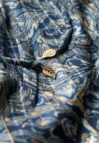 Tissu présentant des motifs complexes en bleu et crème avec des accents dorés ; comprend un pendentif en feuille d'or et une étiquette logo en forme courbée.