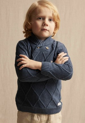 Pull en maille bleu marine avec un motif en losanges, col côtelé et deux boutons décoratifs. Assorti à un pantalon clair.