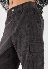 Close-up of dark gray corduroy pants with side cargo pocket and silver button, worn with a patterned top.