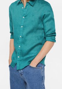Tyrkysová lněná košile s límečkem na knoflíky, jednou náprsní kapsou a vyhrnutými rukávy, kombinovaná s modrými džínami.