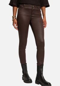 Jeans skinny marroni con una finitura lucida, dotati di tasche anteriori e dettagli borchiati in vita. Abbinati a stivali a caviglia neri e voluminosi.