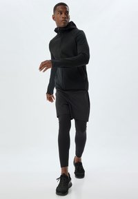Giacca sportiva nera con cappuccio, chiusura con zip e maniche lunghe, abbinata a pantaloni corti neri e leggings. Scarpe da ginnastica nere completano l'outfit.