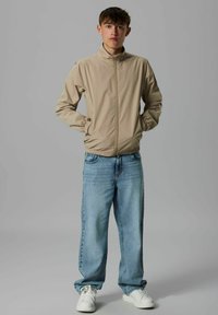Veste légère beige avec un col montant, des poignets élastiques et une fermeture éclair à l'avant, associée à un jean baggy bleu délavé et des baskets blanches.