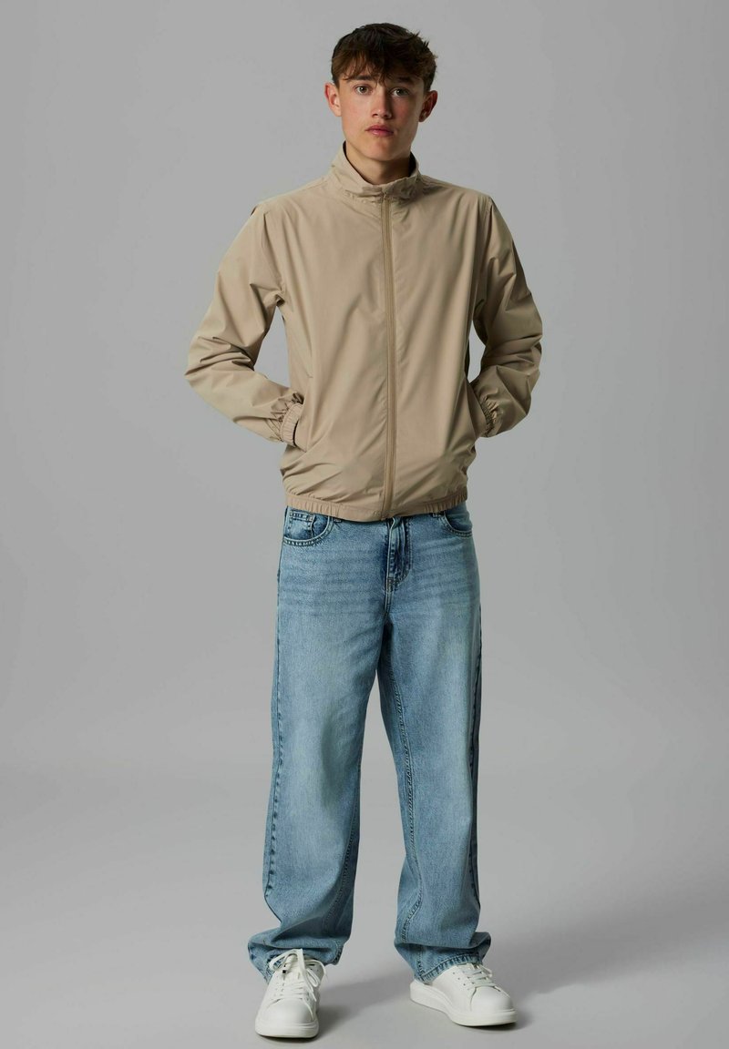 Veste légère beige avec un col montant, des poignets élastiques et une fermeture éclair à l'avant, associée à un jean baggy bleu délavé et des baskets blanches.