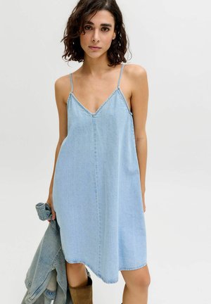 Femme aux cheveux foncés mi-longs portant une robe en jean sans manches bleu clair, tenant une veste en jean, sur un fond uni.