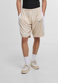 Beige Sportshorts mit weißen Seitenpanelen, elastischem Bund und einem kleinen schwarzen Logo "DEF" am linken Bein. Glatte, leichte Stoffqualität.