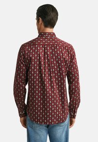 Homme portant une chemise à manches longues bordeaux avec un motif paisley et un jean bleu, se tenant de dos contre un fond uni.