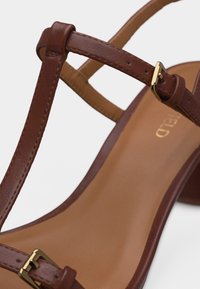 Καφέ δερμάτινο σανδάλι με ρυθμιζόμενους ιμάντες με χρυσή αγκράφα και ταμπά μαλακό πάτο σε λευκό φόντο.