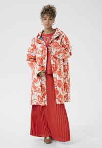 Dážďový plášť s kvetinovým vzorom v oranžovej a krémovej farbe, s kapucňou a zapínaním na cvoky, doplnený skladanou červenou sukňou a hnedými topánkami.