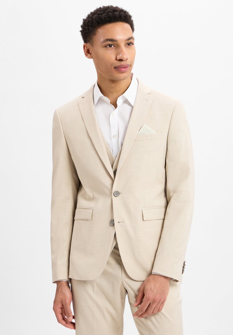 Lichtbeige pak met een enkelzijdig jasje, geknipte revers en een borstzak. Wit overhemd eronder en bijpassende broek.