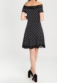 Svart prickig klänning med off-the-shoulder spetskrage och utsvängd kjol. Har en spetskant vid fållen. Matchas med svarta klackskor.