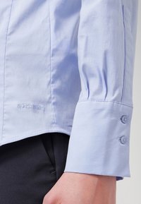 Camicia azzurra chiara con una texture liscia, caratterizzata da un design con maniche a sbuffo, dettagli con bottone ai polsini e un logo discreto all'orlo.