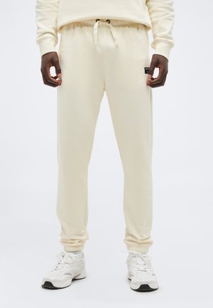 Άνδρας που φοράει ανοιχτό μπεζ αθλητικό παντελόνι με ελαστική μέση και κορδόνι, σε συνδυασμό με λευκά αθλητικά παπούτσια.