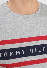 Camiseta de algodón gris con cuello redondo, que presenta llamativas rayas horizontales en rojo y azul marino, con texto blanco que dice "TOMMY HILFIGER."