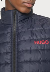 Marineblaues gepolstertes Weste mit hohem Kragen, seitlichem Reißverschluss und rotem "HUGO"-Logo auf der Brust. Glatte, glänzende Textur mit gestepptem Design.
