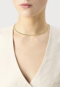 Zlatá reťazová choker náhrdelník s plochým dizajnom, obsahujúca prepojené články; vystavená na neutrálnom odeve.