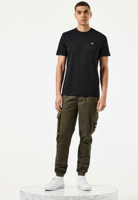 Zwarte katoenen t-shirt met een klein logo op de borst, gecombineerd met olijfgroene cargobroeken met zijzakken en elastische boorden, gedragen met witte sneakers.