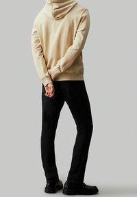 Sudadera beige con capucha y puños acanalados, combinada con jeans slim fit negros y calzado negro. Textura suave, diseño casual.