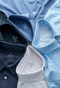 Samling av polo shirts i olika nyanser av blått, med enkla och mönstrade designer, krage och knappar. Etiketter synliga.