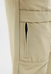 Beige hlače imaju uski kroj s džepom na zatvarač i glatkom teksturom tkanine. Detalji šivanja su vidljivi duž šavova.