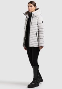 Giacca bianca imbottita con colletto alto, zip frontale e tasche laterali; abbinata a leggings neri e stivali neri alti fino al ginocchio.
