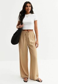 Camiseta blanca corta, pantalones beige de pierna ancha con lazo en la cintura, bolsa tejida negra, sandalias planas. Tela ligera y casual, textura suave.