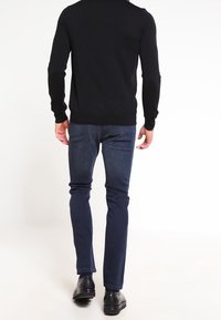 Suéter negro de hombre con cuello redondo, combinado con jeans azul oscuro y zapatos negros, visto desde atrás. Textura suave y diseño ajustado.