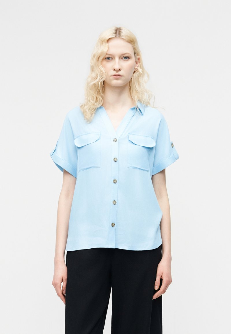 Γυναίκα με κυματιστά ξανθά μαλλιά, φοράει ανοιχτό μπλε πουκάμισο με κοντά μανίκια και μαύρο παντελόνι, στέκεται μπροστά σε απλό φωτεινό φόντο.