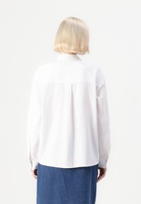 Camicia bianca a maniche lunghe con colletto, caratterizzata da una piega sul retro, realizzata in tessuto liscio. Modello abbinato a una gonna di jeans.