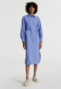 Helles blaues, langärmliges Hemdkleid mit gebundenem Taillenzug, seitlichen Schlitzen und strukturiertem Stoff. Kombiniert mit beigen Plateauschuhen.
