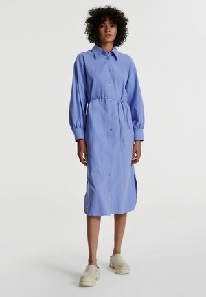 Helles blaues, langärmliges Hemdkleid mit gebundenem Taillenzug, seitlichen Schlitzen und strukturiertem Stoff. Kombiniert mit beigen Plateauschuhen.