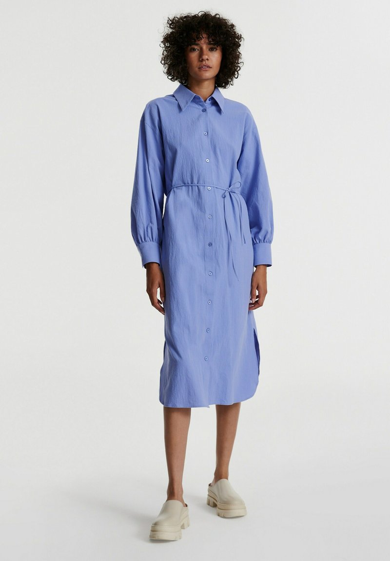Helles blaues, langärmliges Hemdkleid mit gebundenem Taillenzug, seitlichen Schlitzen und strukturiertem Stoff. Kombiniert mit beigen Plateauschuhen.