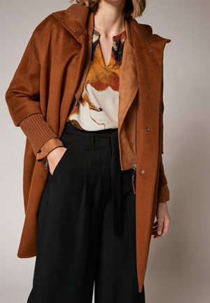 Femme portant un manteau marron rouille sur un haut à motifs avec un pantalon noir taille haute et jambes larges, debout avec une main dans la poche sur fond uni.